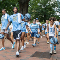A group of kids and young adults bouncing basketballs on a college campus.