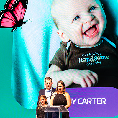 A family giving a speech in remembrance of their son Carter on a stage with a picture of Carter in the background.