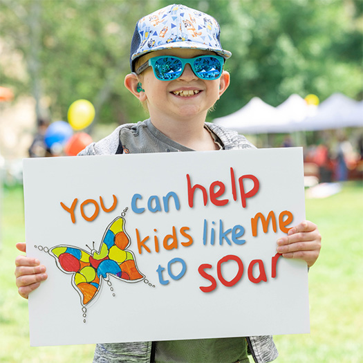A kid with a hat and sunglasses on holding a sign that reads "you can help kids like me to soar"