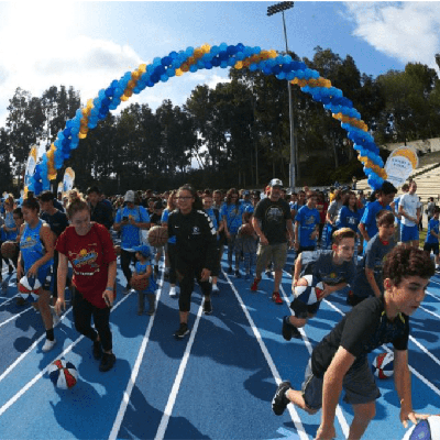 A group of children dribbling basketballs on a college track surface with a balloon arch in the background.